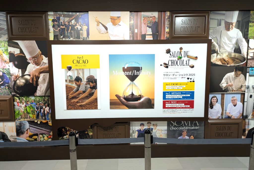 開場の壁面装飾。サロン・デュ・ショコラのポスター、チョコレート職人、カカオ生産者の写真が並ぶ。写真のあいまにチョコレート形のパネルがあり、そこに「SALON DU CHOCOLAT」と刻まれており、その右上には商標登録を示すアールマークが刻まれている。